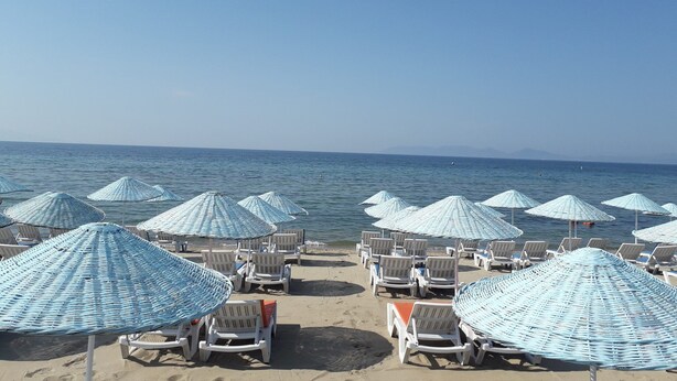 A beach with rows of white lounge chairs and blue umbrellas. - Nil Hotel (Ayvalik)
