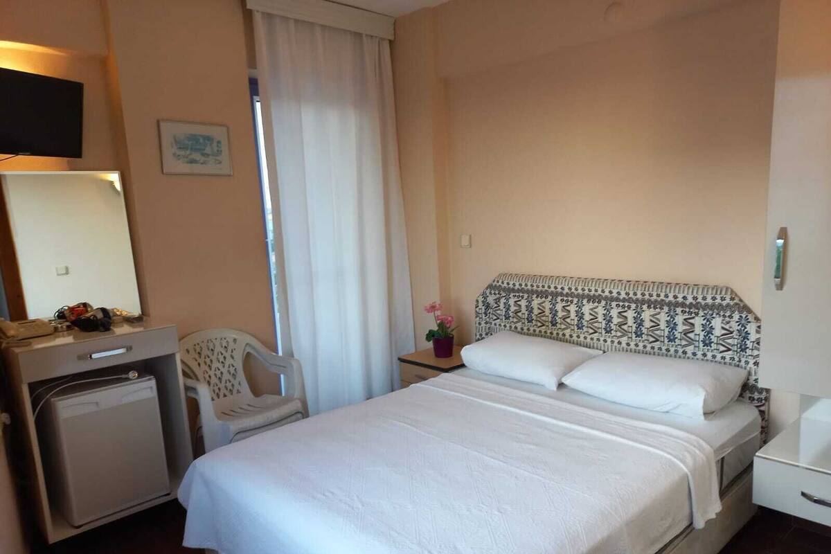 A hotel room with a bed, a desk with a computer, a chair, a small table with a flower, and a window with curtains.