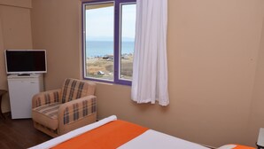 A room with a bed, a chair, a television, and a window with a view of the beach. - Nil Hotel (Ayvalik)