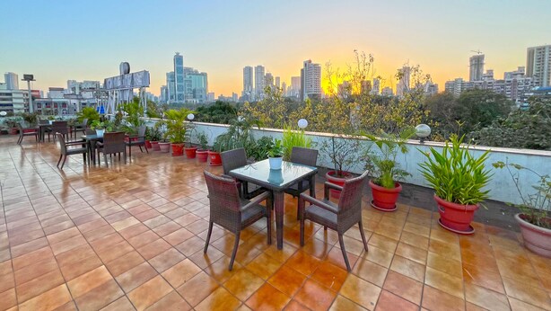Una terraza en la azotea con mesas y sillas, plantas en macetas y un horizonte urbano al fondo.