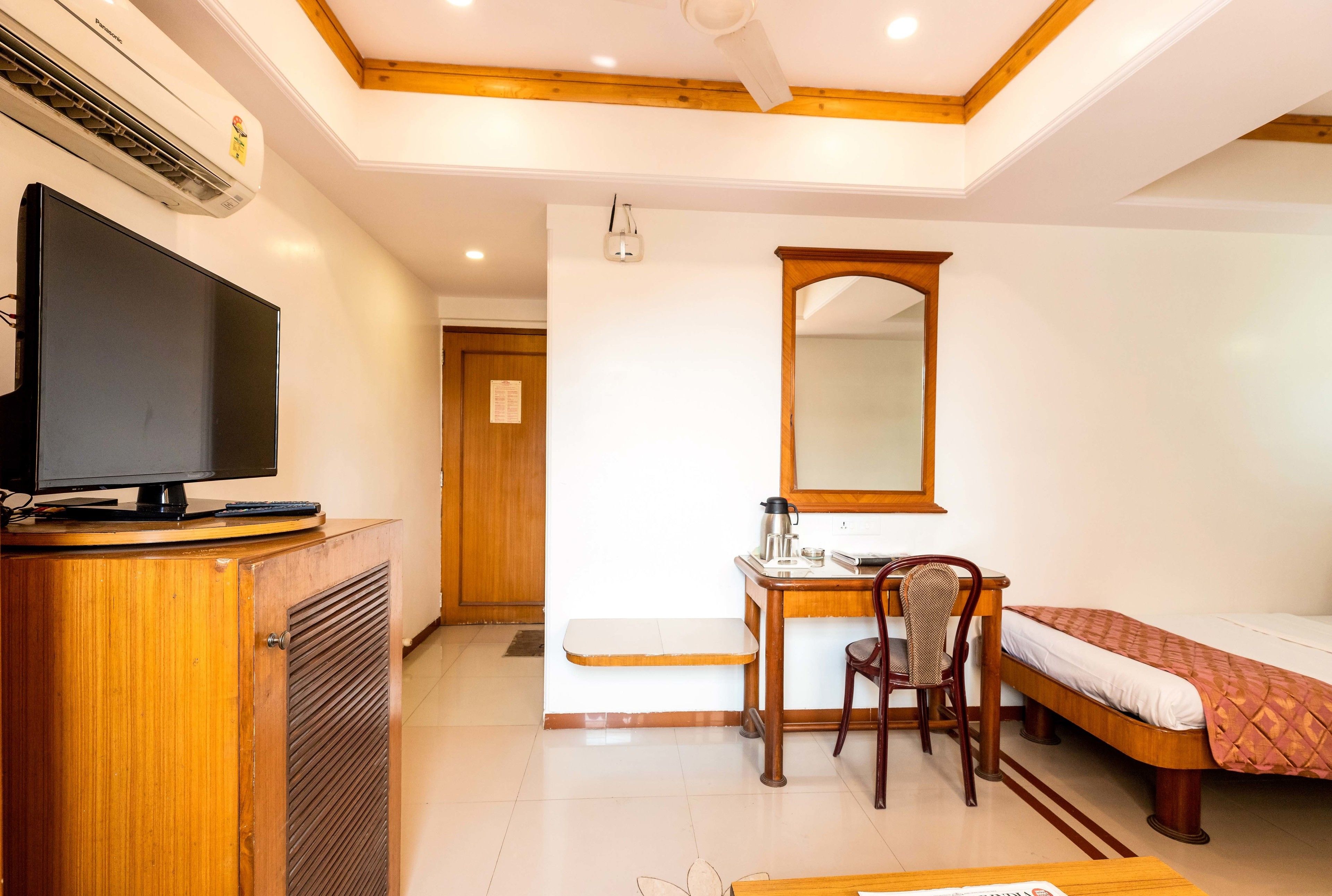 A hotel room with a wooden desk, a chair, a bed, a television on a cabinet, and a ceiling fan.