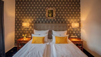Ein Doppelbett mit weißen Leinen und zwei gelben Kissen, an beiden Seiten Nachttische mit Lampen, in einem Zimmer mit gemustertem Wandpapier und einem gerahmten Bild über dem Bett.