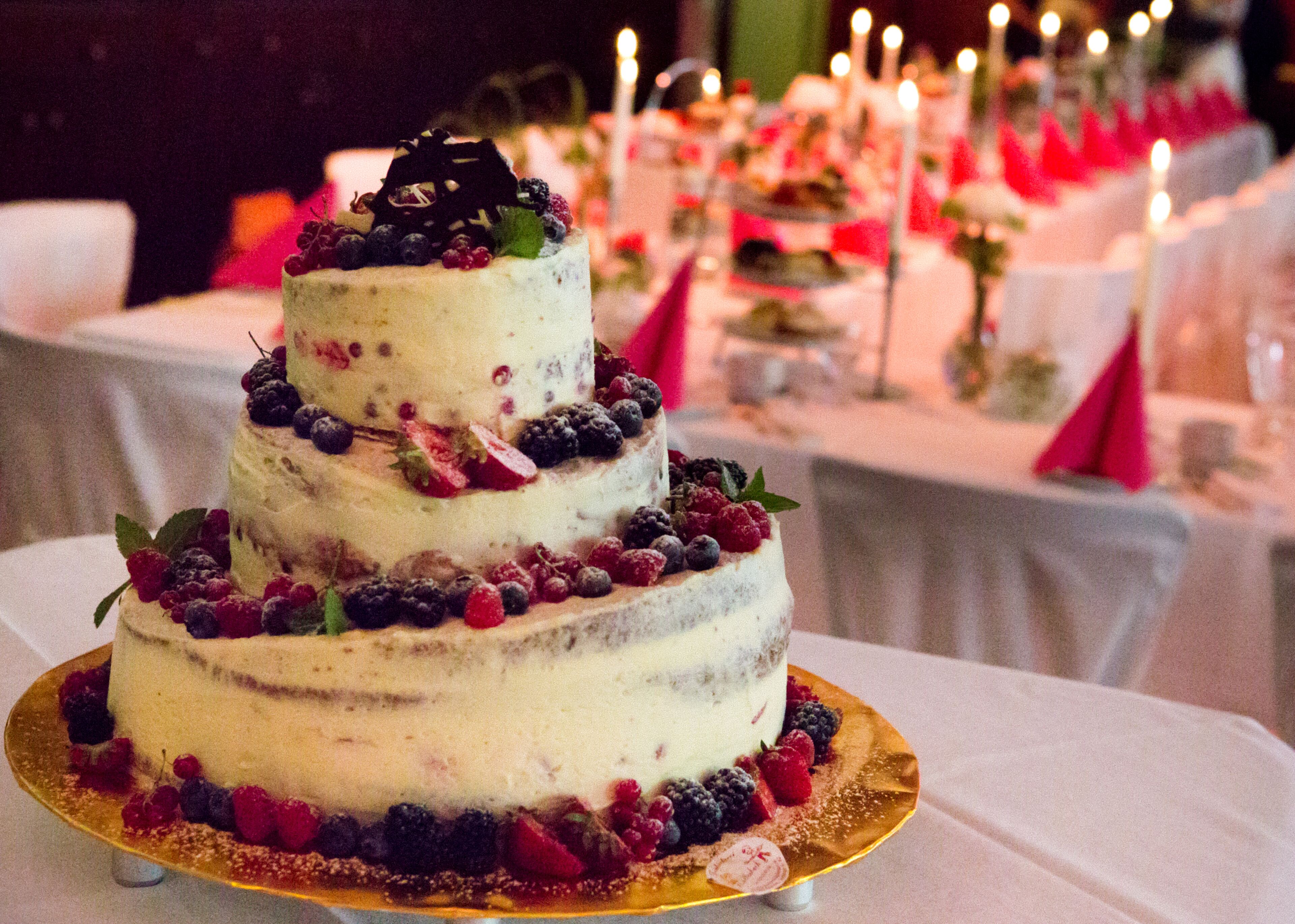 A three-tiered cake with fruit decorations on a golden cake stand, placed on a table with white tablecloths and candles in the background.