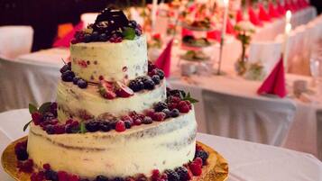 A three-tiered cake with fruit decorations on a golden cake stand, placed on a table with white tablecloths and candles in the background.