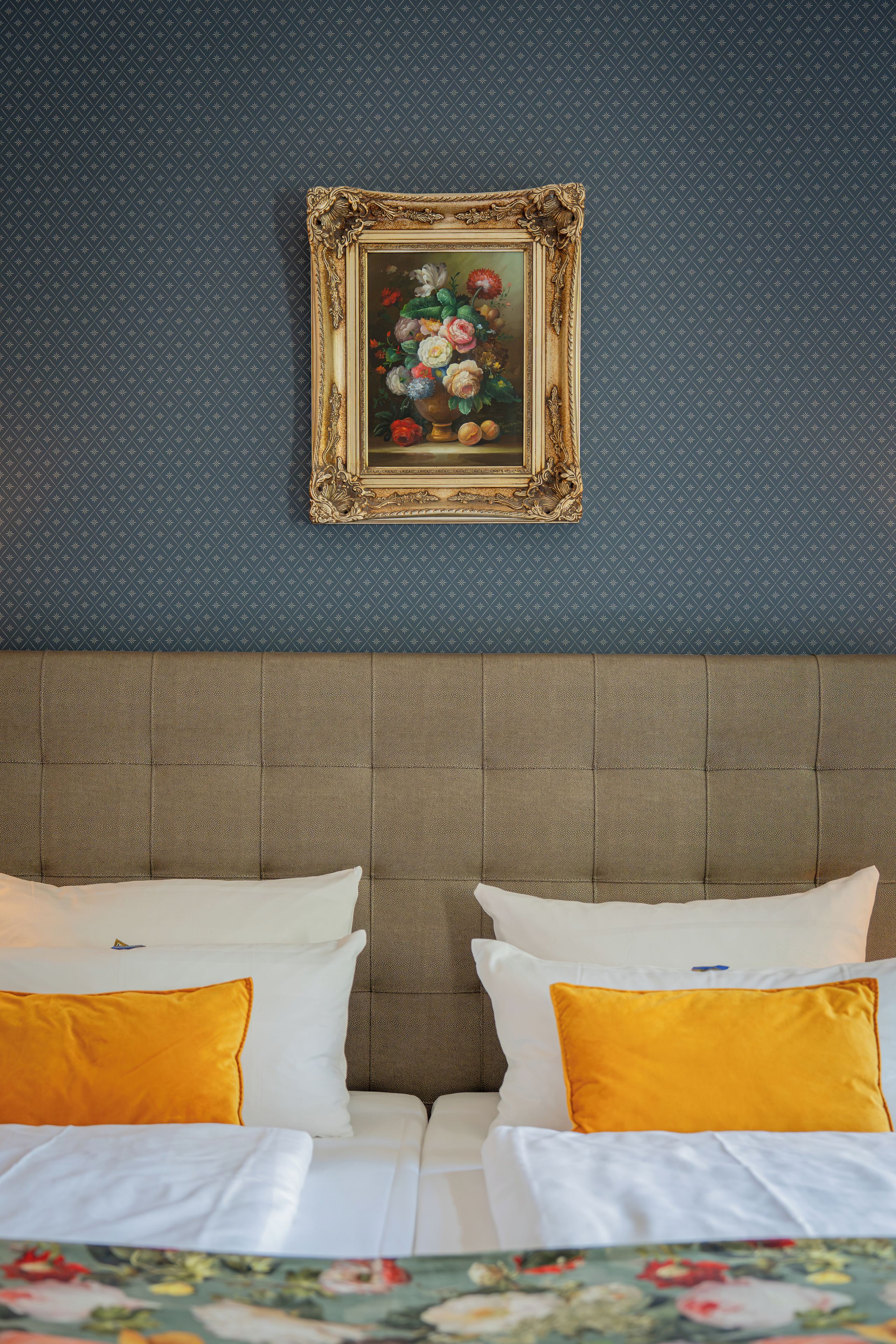 A hotel room with a headboard, two white pillows, and a framed floral artwork on the wall.