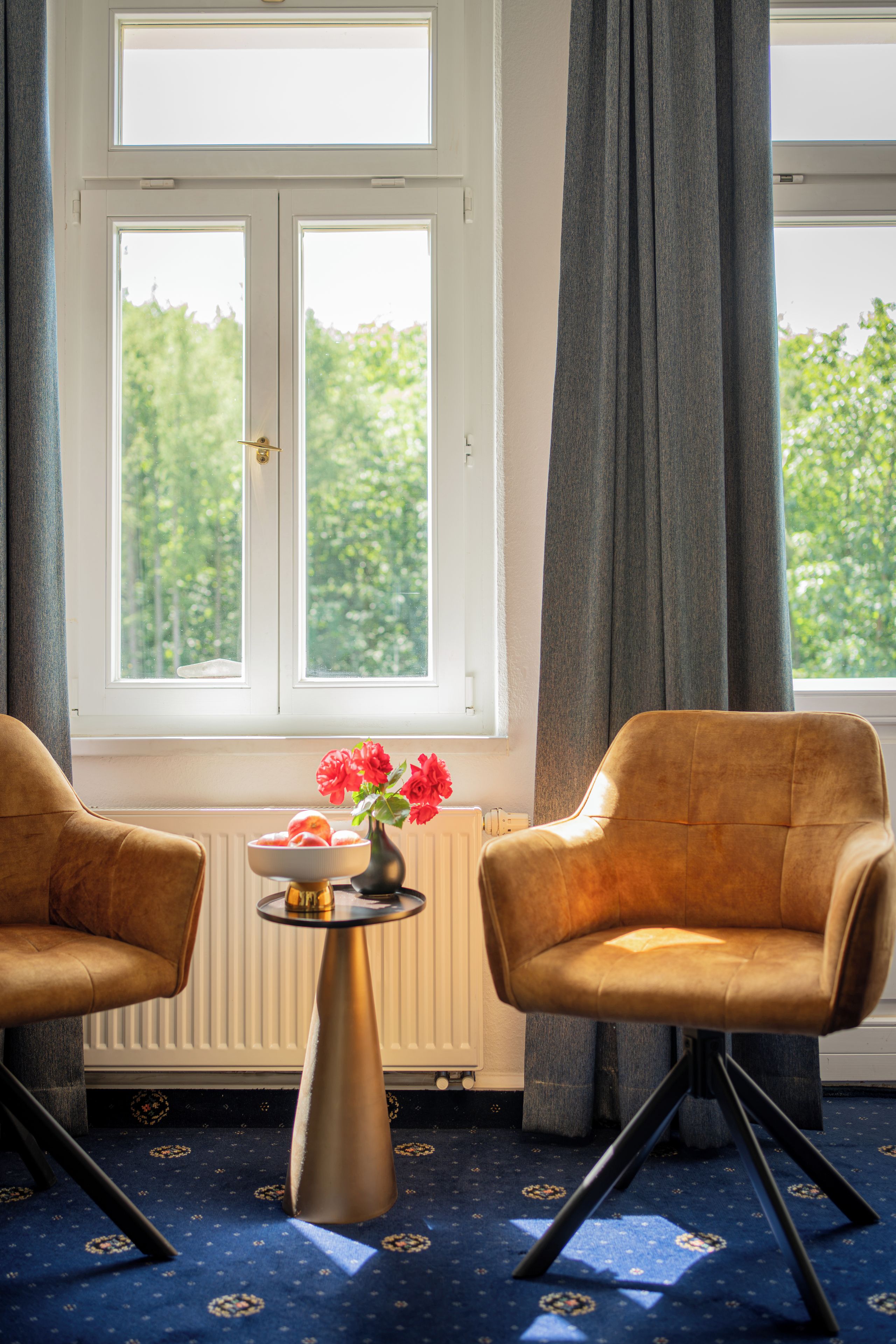 Een kamer met twee fauteuils, een bijzettafeltje met een bloemenvaas en een raam met gordijnen.