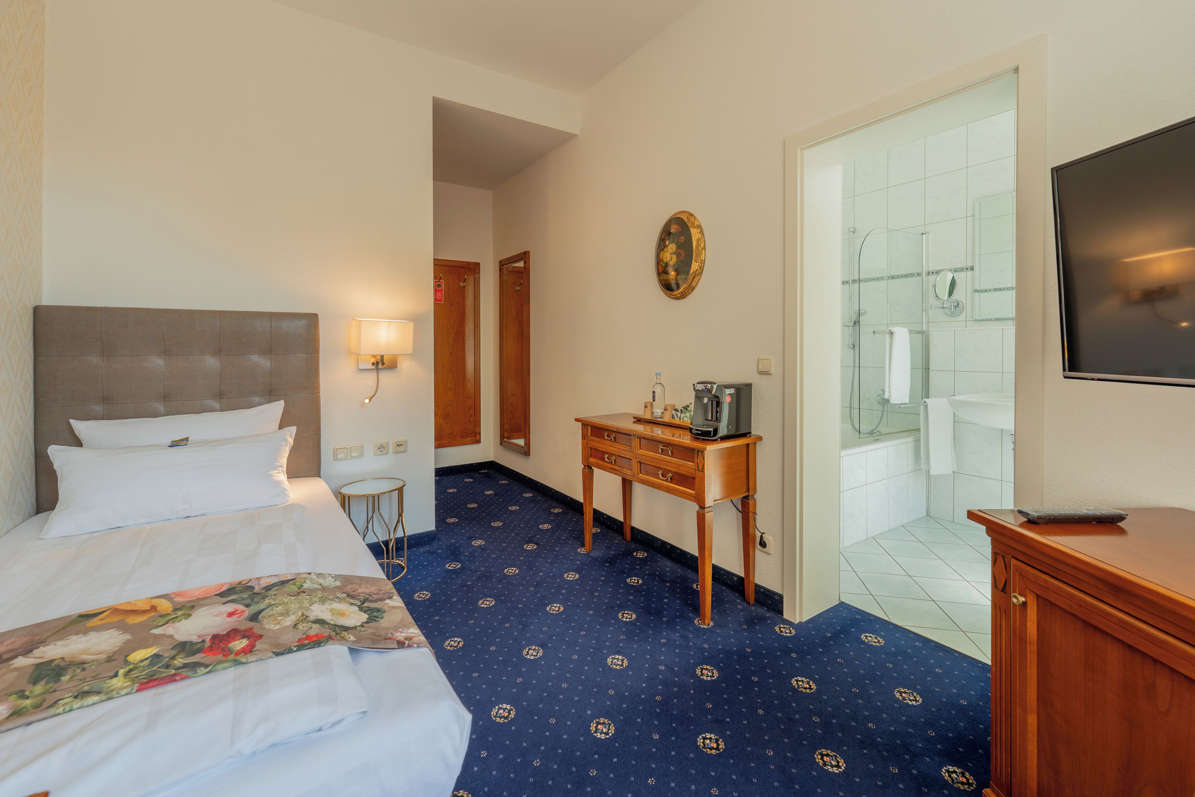 A hotel room with a bed, a desk, a chair, a television, and a bathroom visible through an open door.
