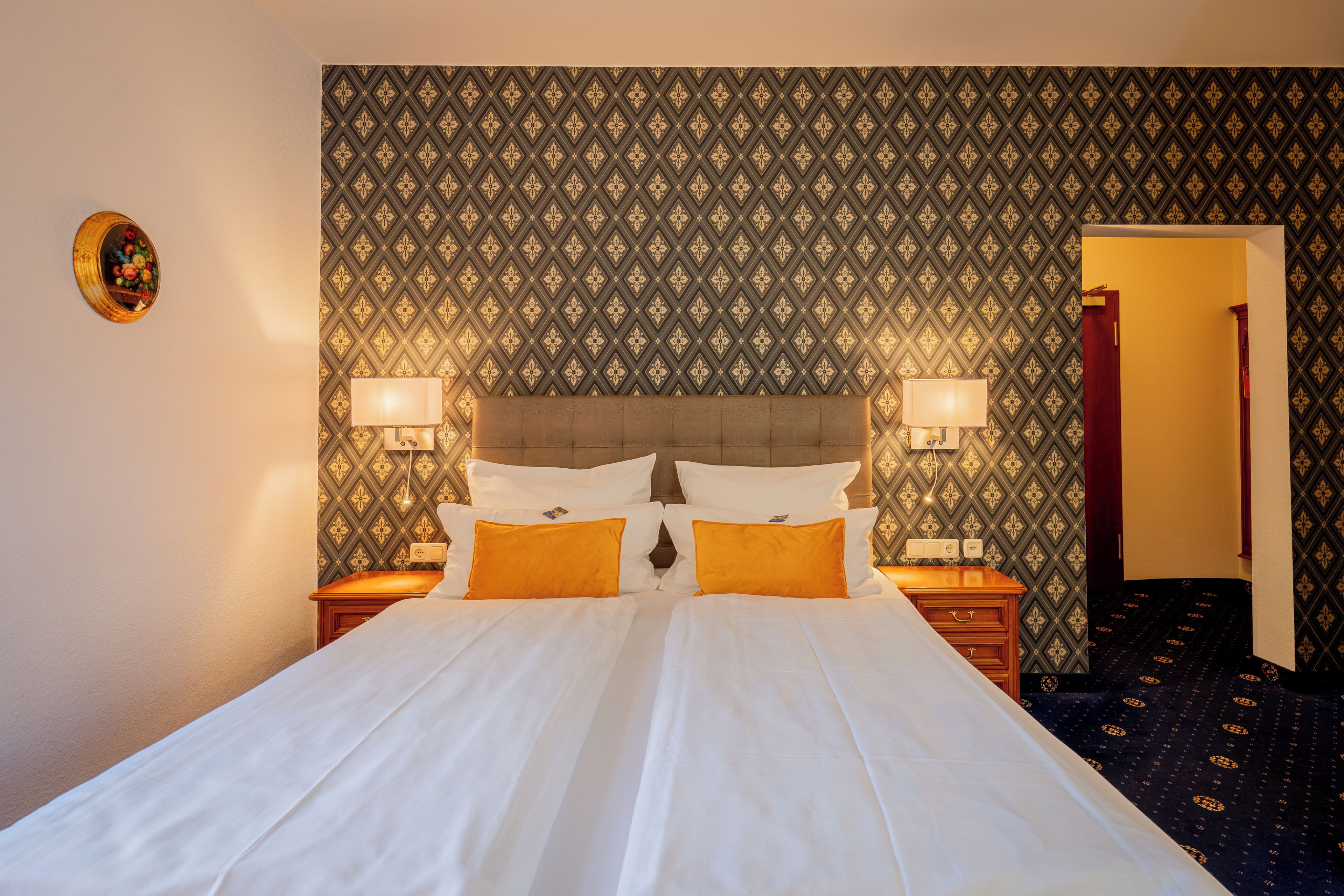 Una habitación de hotel con papel tapiz estampado, una cama con ropa blanca y dos almohadas, lámparas de noche y una mesita de madera.