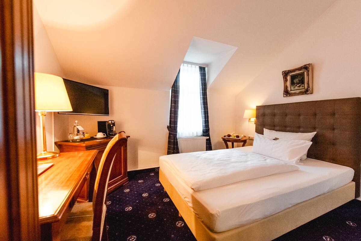 A hotel room with a bed, a desk, a chair, a television, a window with curtains, and a picture on the wall.