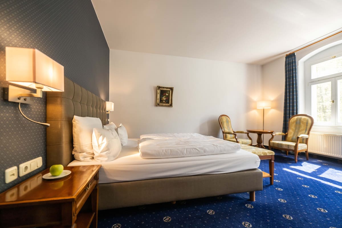 A hotel room with a bed, bedside table, lamp, two chairs, a small table, and a picture on the wall.