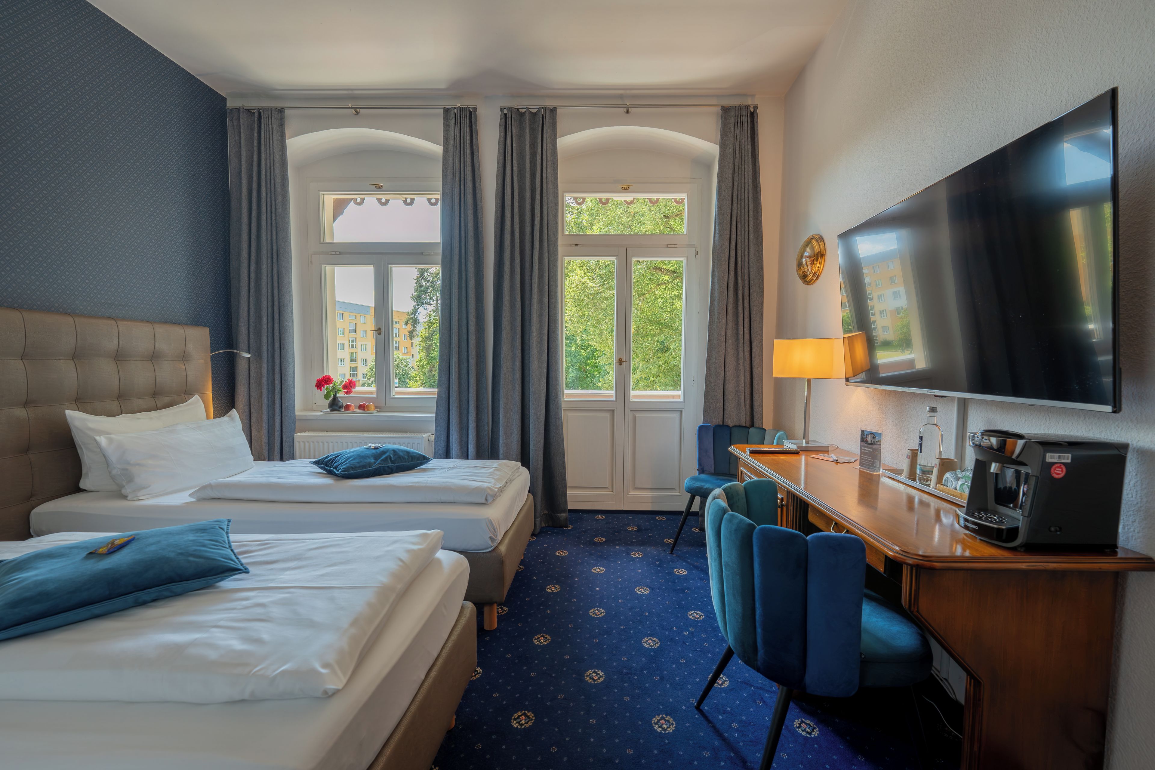 Habitación de hotel con dos camas, un escritorio con cafetera, televisor y vista a zonas verdes.