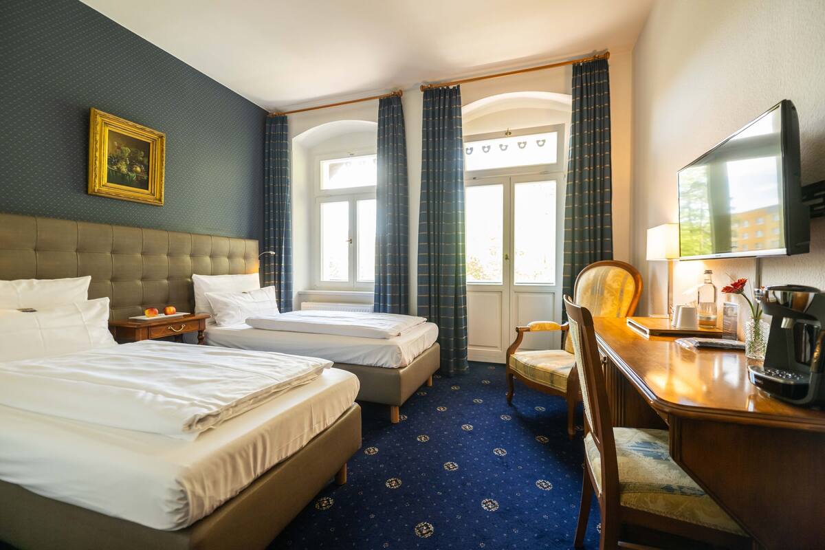 A hotel room with two beds, a wooden desk, a chair, a TV, and a window with curtains.