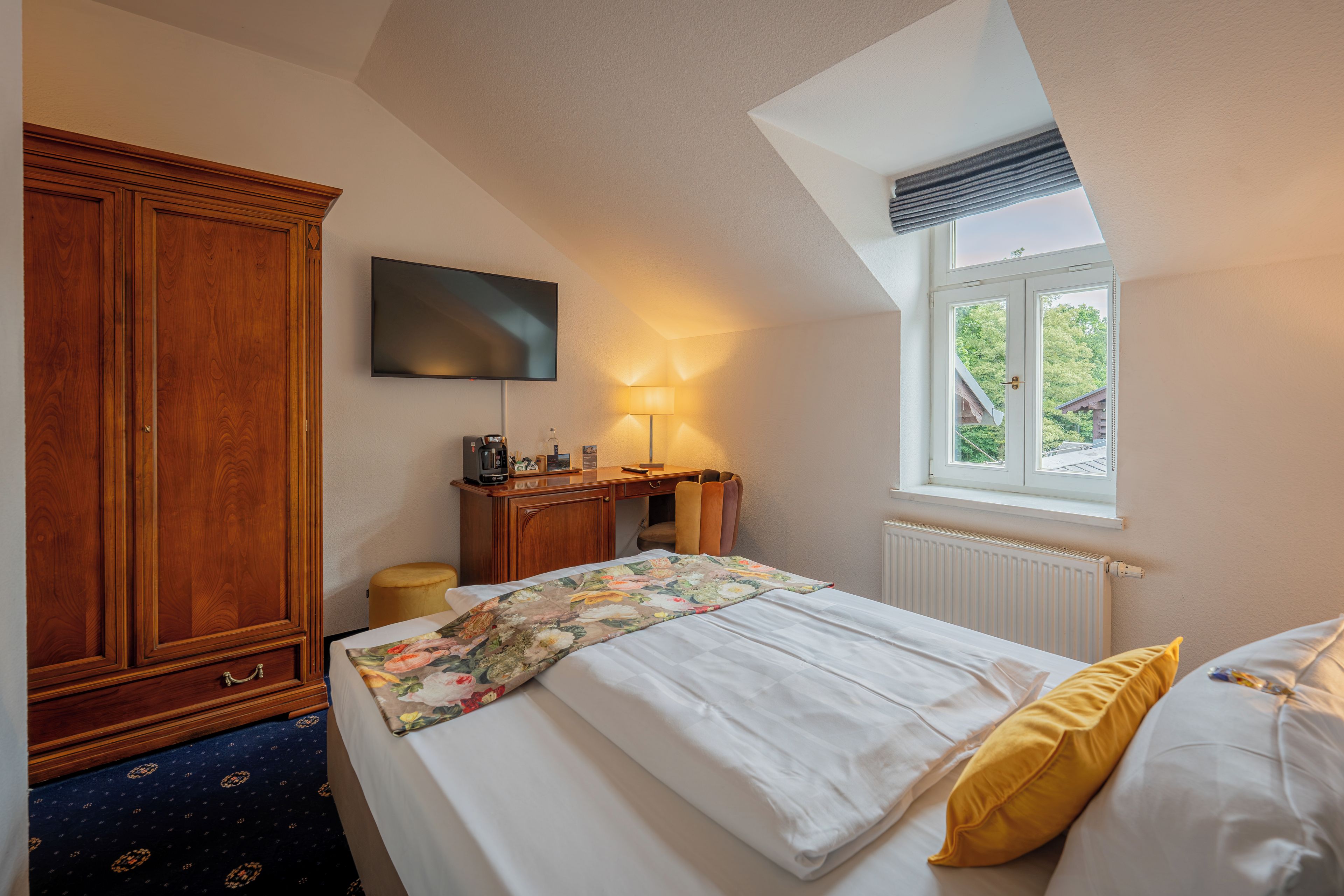 A bedroom with a bed, a wardrobe, a desk with a lamp, a television, and a window with a view of greenery.