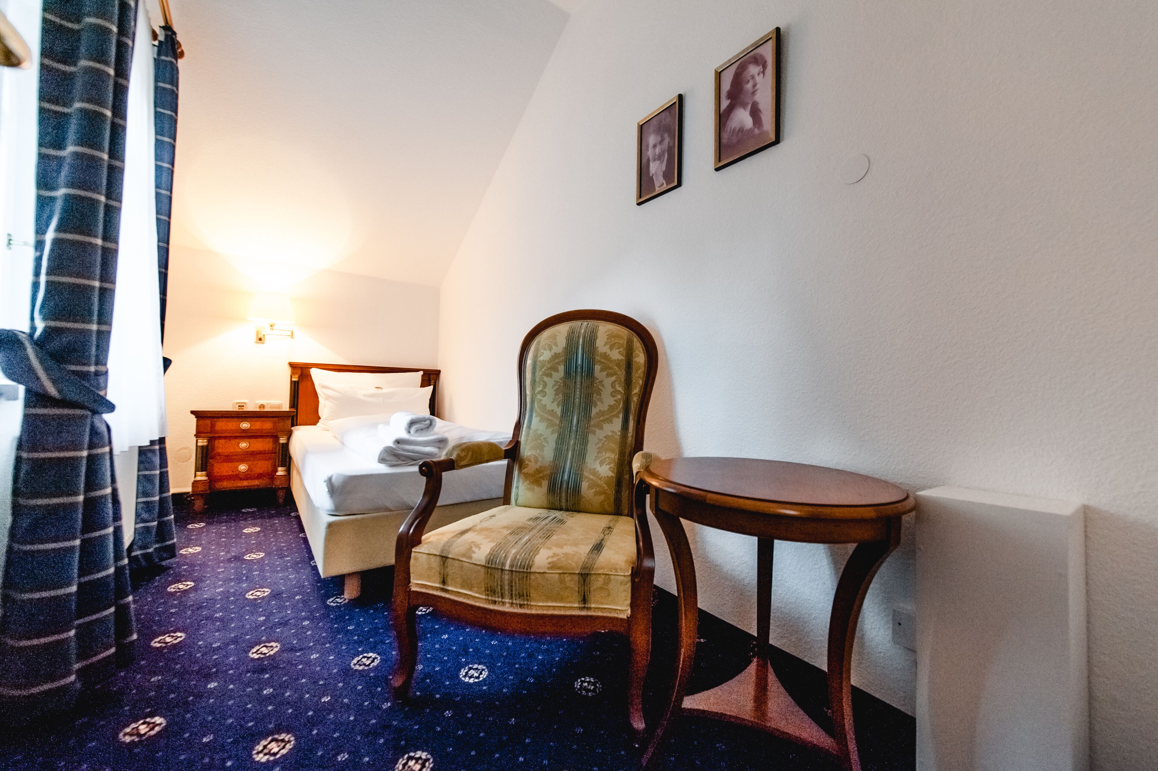 A hotel room with a bed, a chair, a small table, and a nightstand with a lamp.