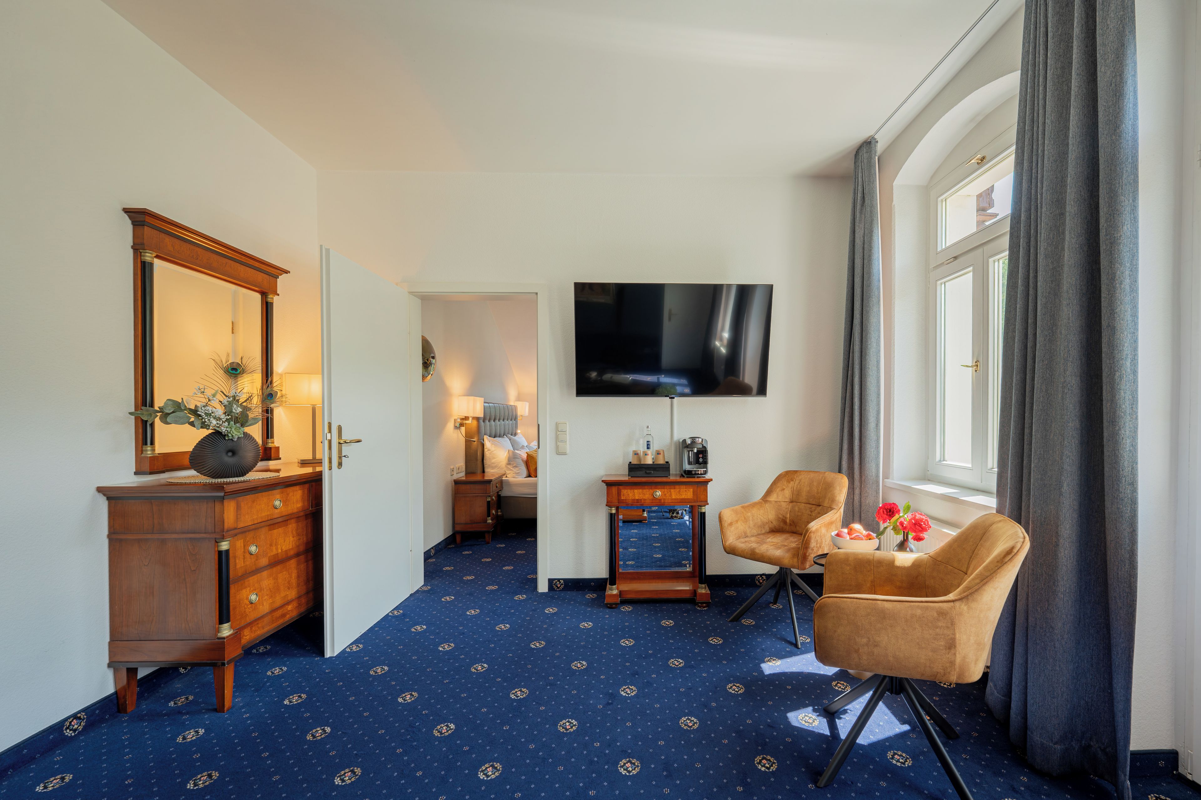 Habitación de hotel con un espejo grande, televisión, escritorio con lámpara, silla y ventana con cortinas.