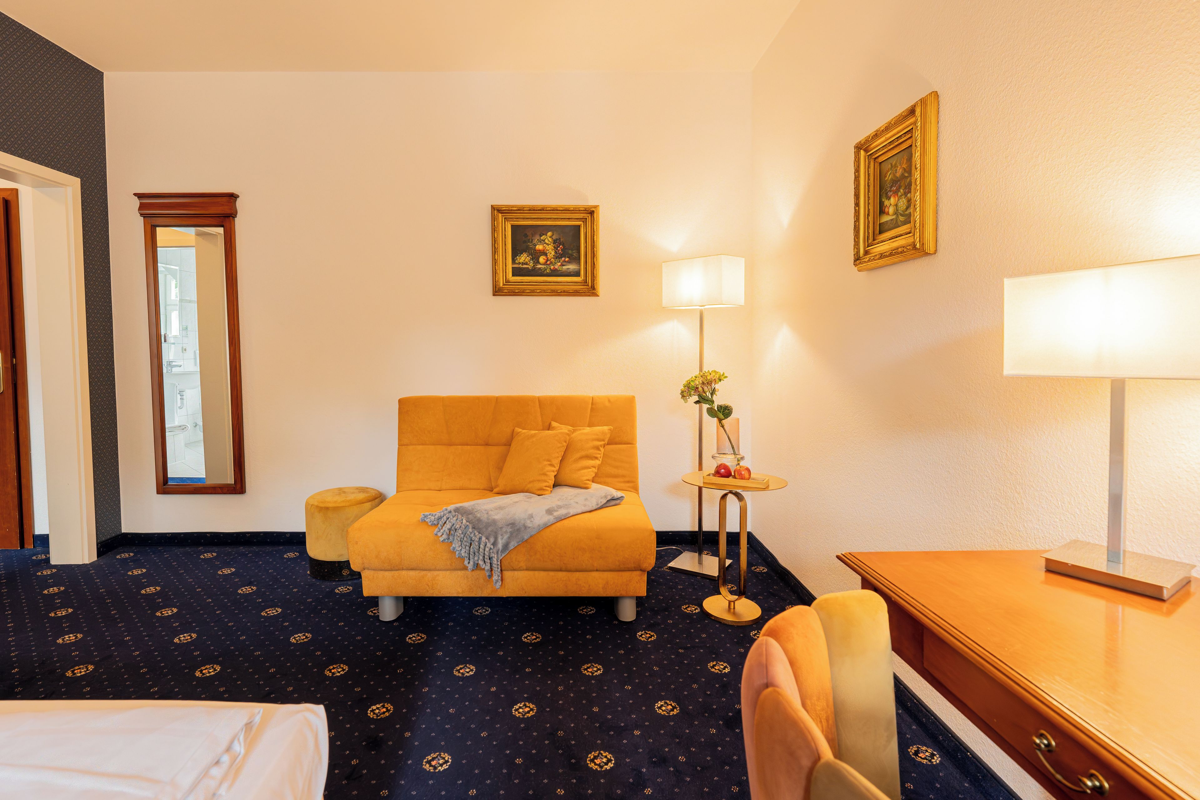 Una habitación de hotel con cama, sofá, escritorio, lámpara y un cuadro en la pared.