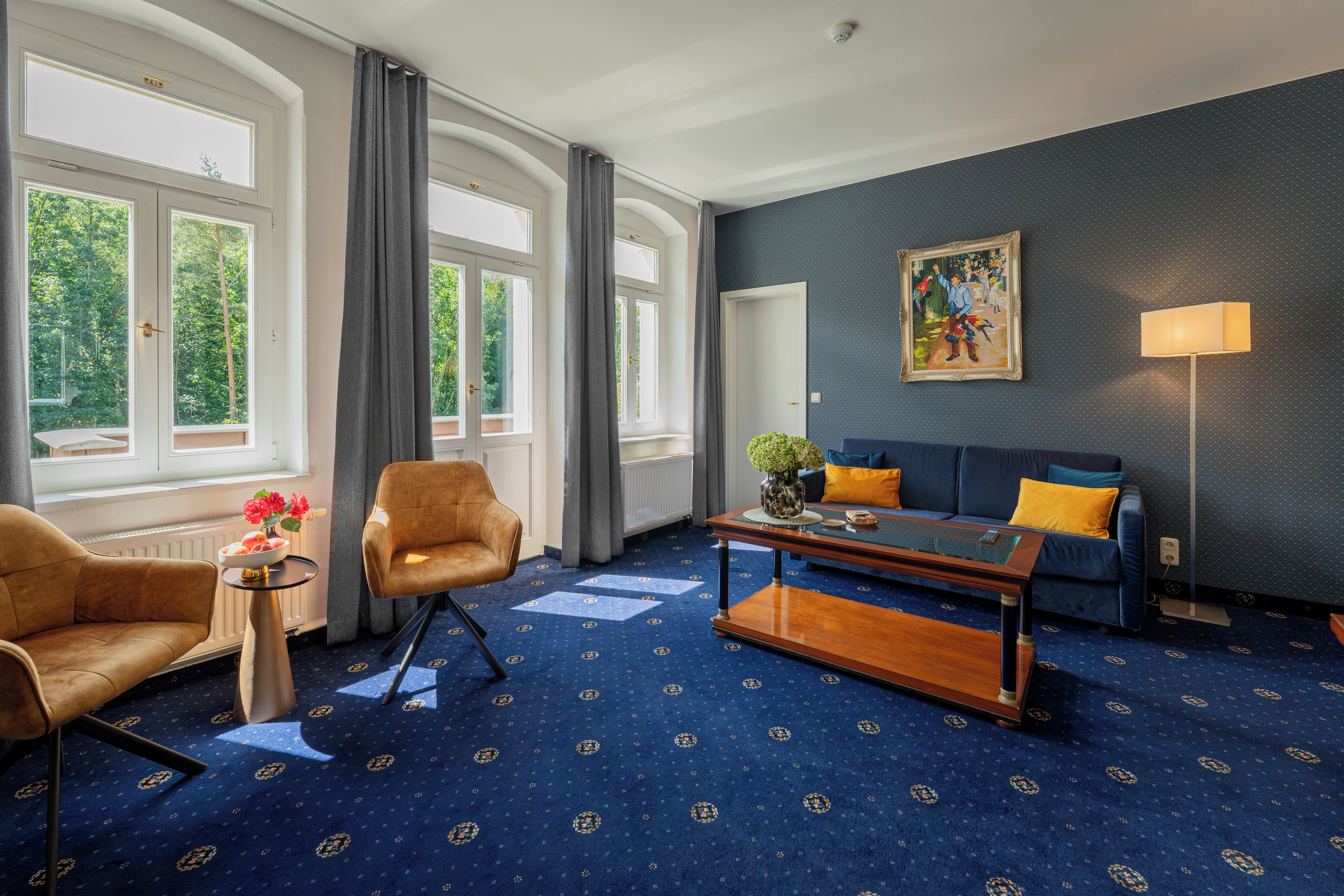Una habitación de hotel con alfombra azul, un sofá, una silla, una mesa de centro y un cuadro en la pared.