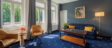 Una habitación de hotel con alfombra azul, un sofá, una silla, una mesa de centro y un cuadro en la pared.