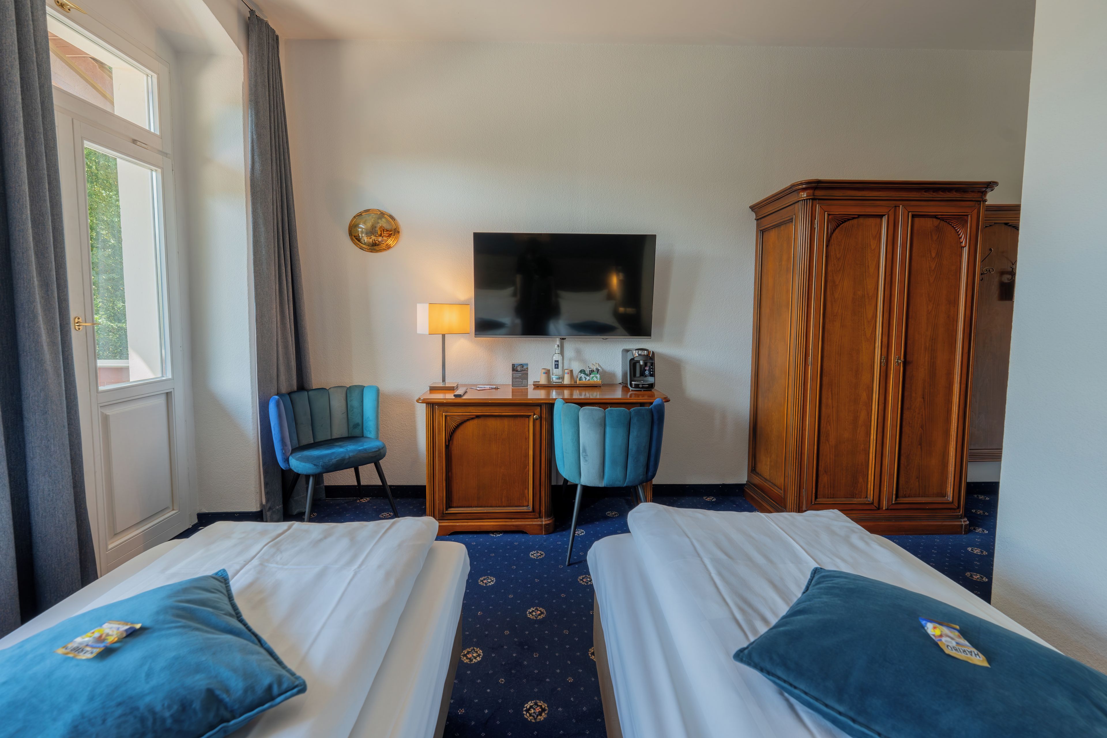 Une chambre d’hôtel avec deux lits, un bureau avec une chaise, une télévision, une armoire et une fenêtre avec des rideaux.