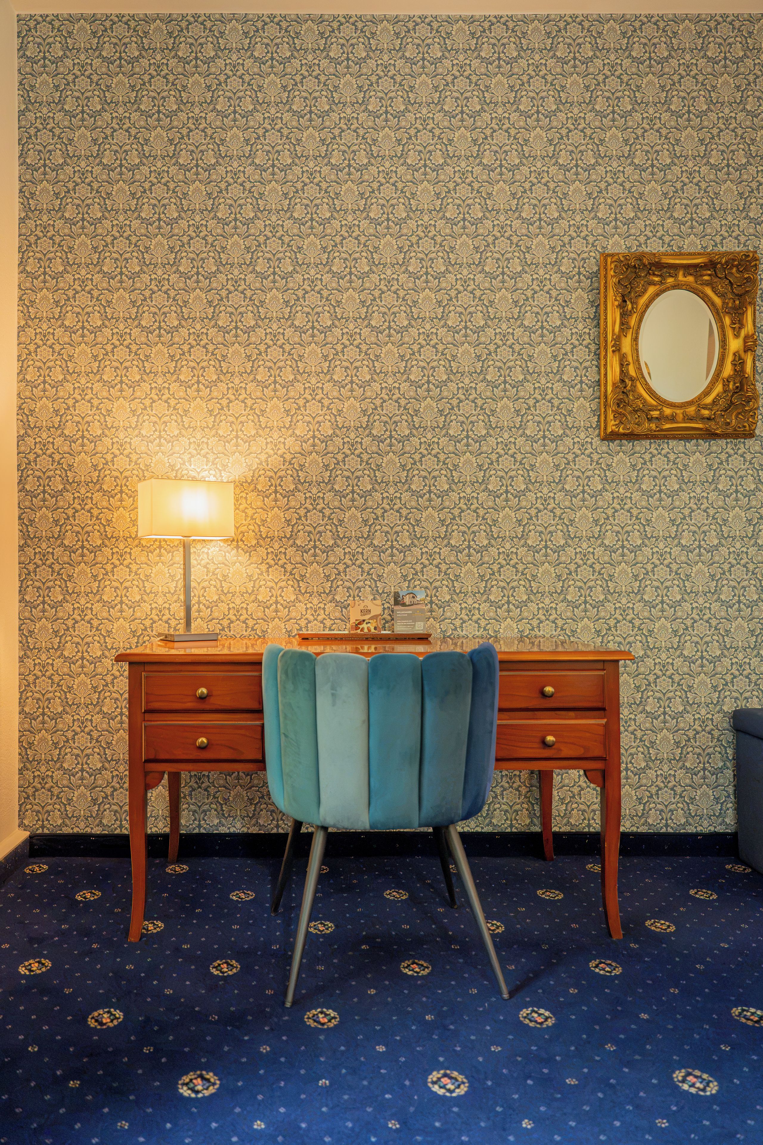 A room with a patterned wallpaper, a desk with drawers, a blue chair, a lamp, and a mirror on the wall.
