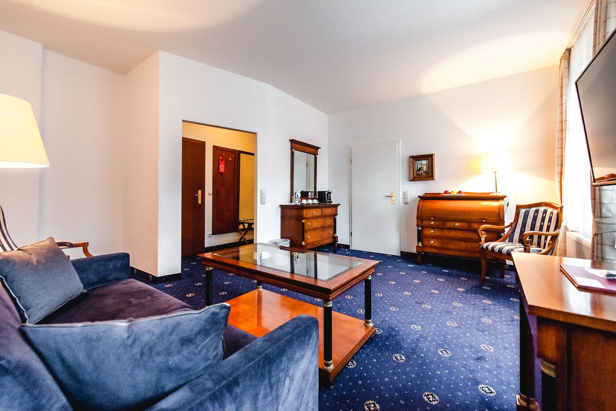A hotel room with a blue patterned carpet, a glass-top coffee table, a blue sofa, a wooden dresser, and a striped armchair.