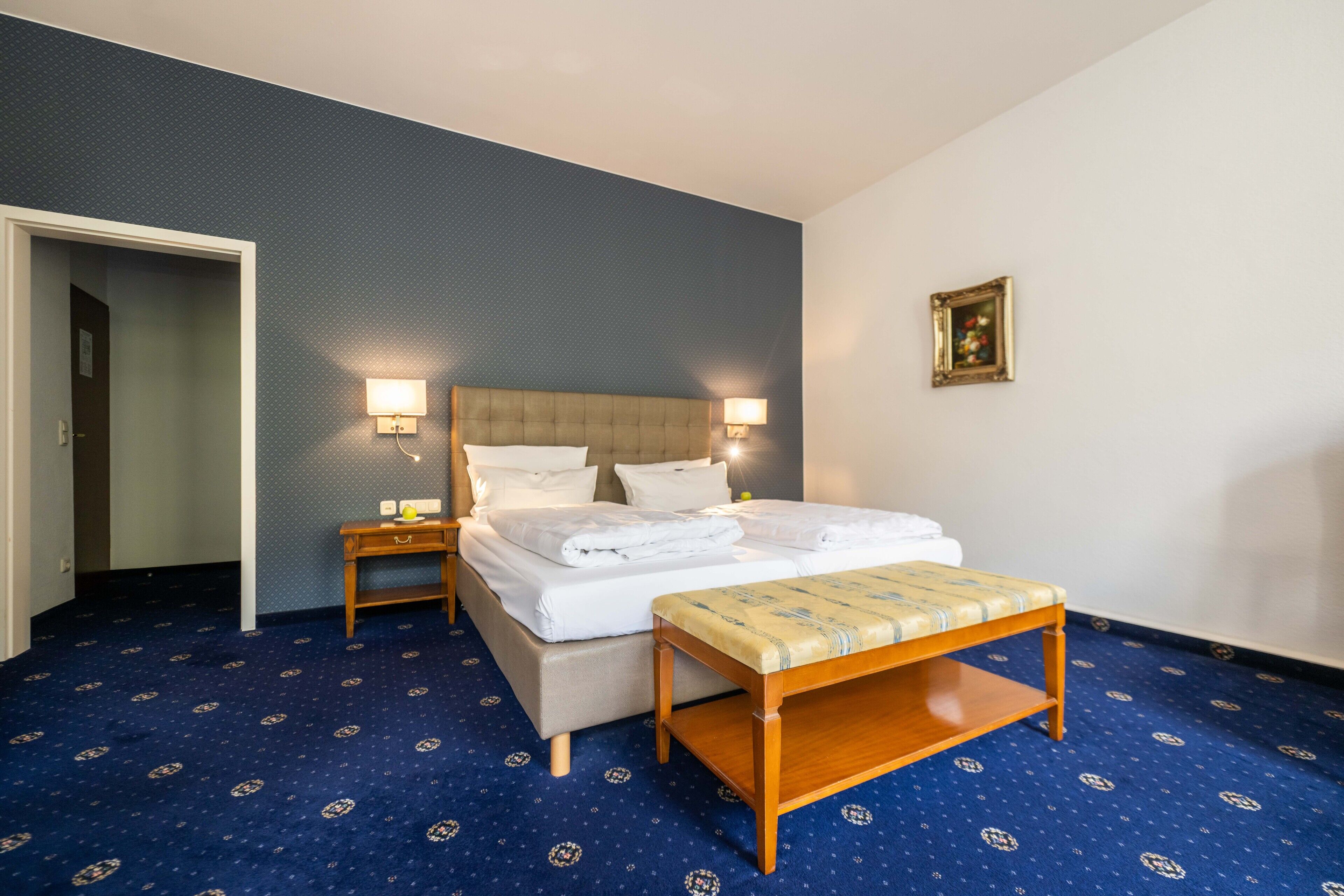 A hotel room with a large bed, bedside tables, a bench, and a framed picture on the wall.