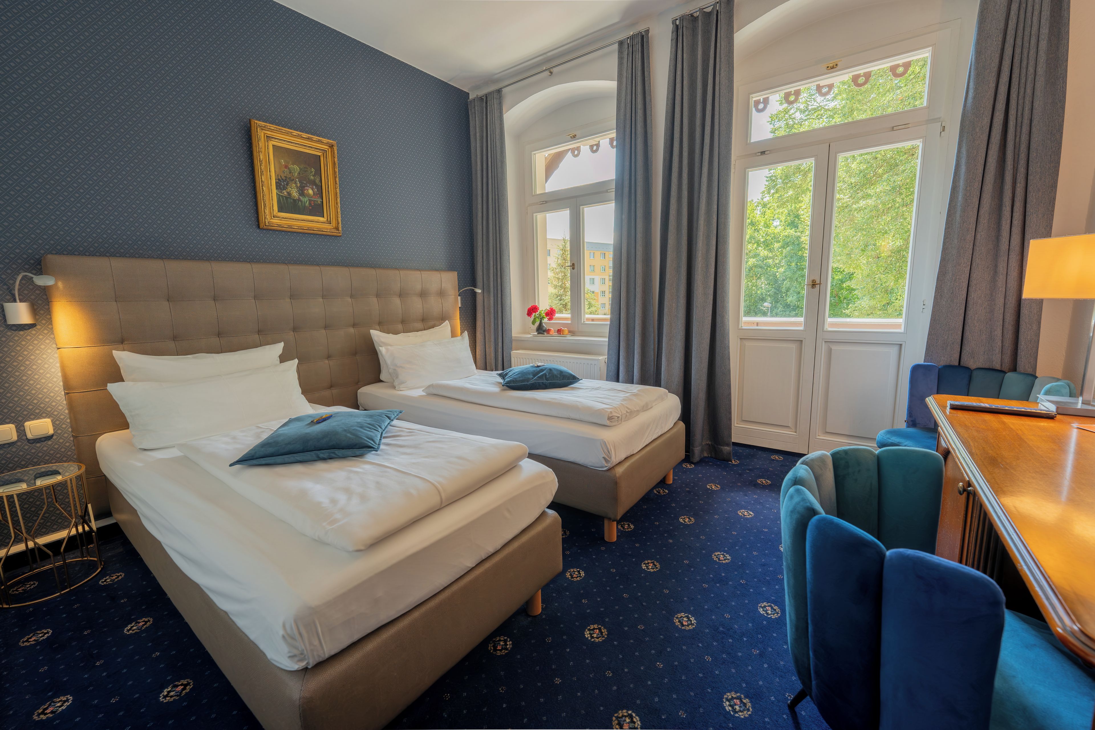 Ein Hotelzimmer mit zwei Betten, einem Schreibtisch, einem Stuhl, einem fenster mit Vorhängen, einem Bild an der Wand und einem gemusterten Teppich.