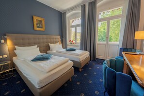 A hotel room with two beds, a desk, a chair, a window with curtains, a painting on the wall, and a patterned carpet. - Historische Spitzgrundmühle Coswig (Coswig)