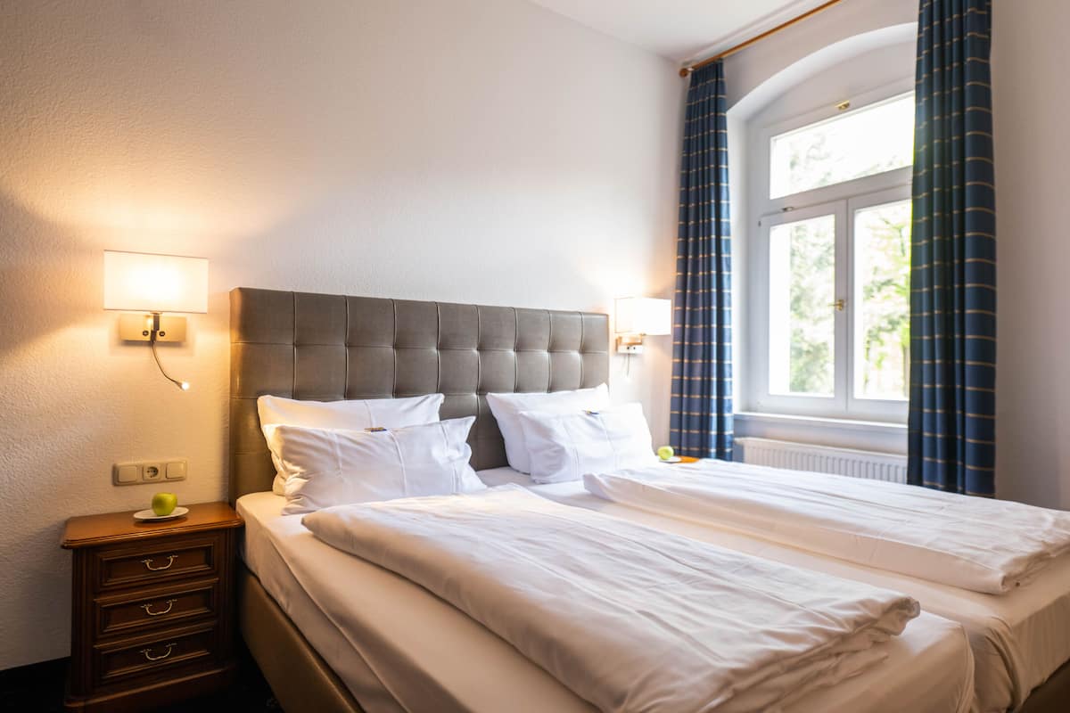 A hotel room with a double bed, a nightstand with drawers, a lamp, and a window with curtains.