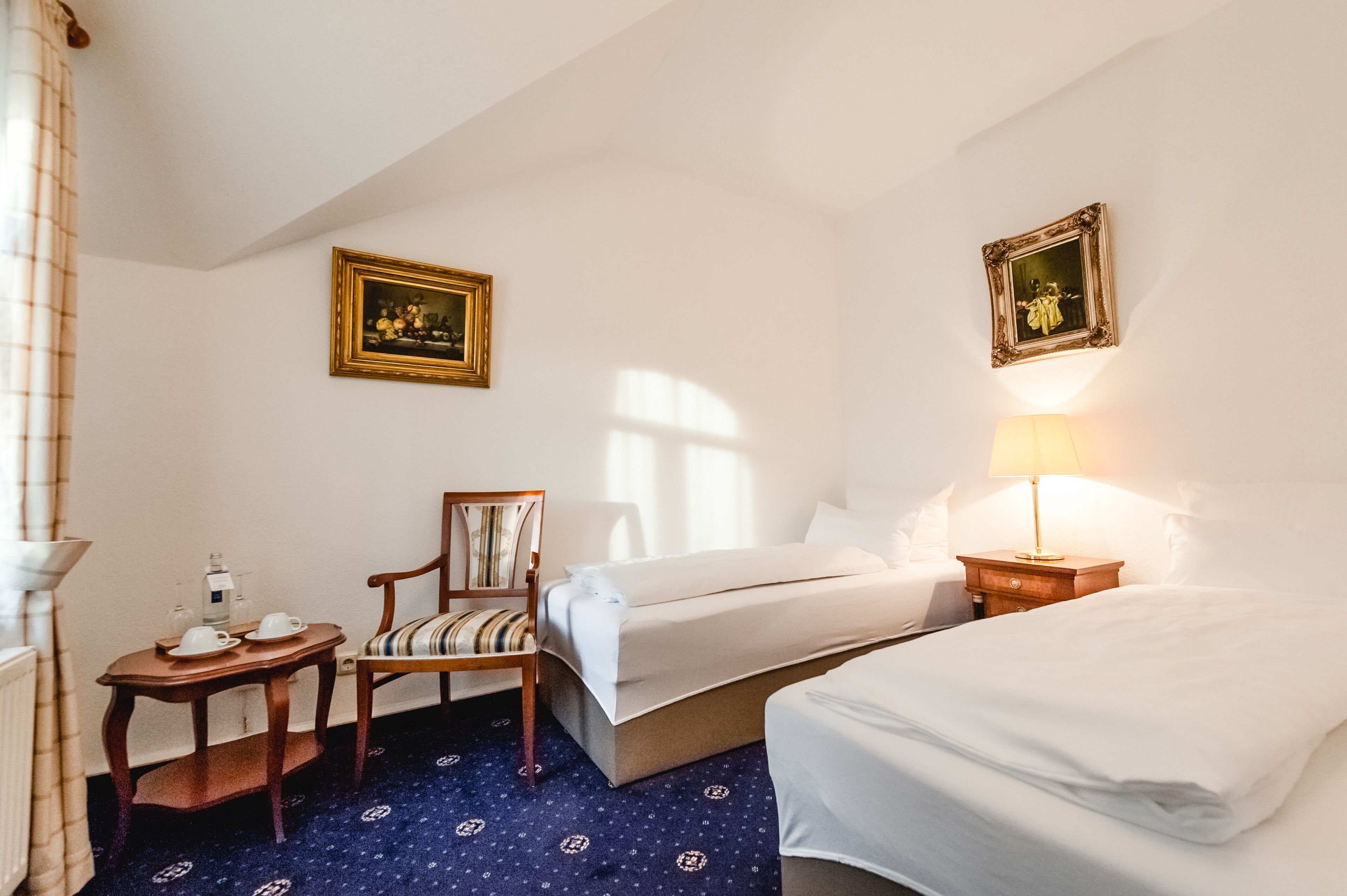 A hotel room with two beds, a chair, a small table with a lamp, and two framed pictures on the walls.