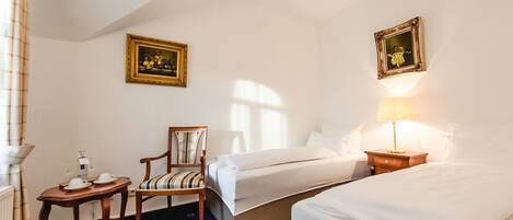 A hotel room with two beds, a chair, a small table with a lamp, and two framed pictures on the walls.