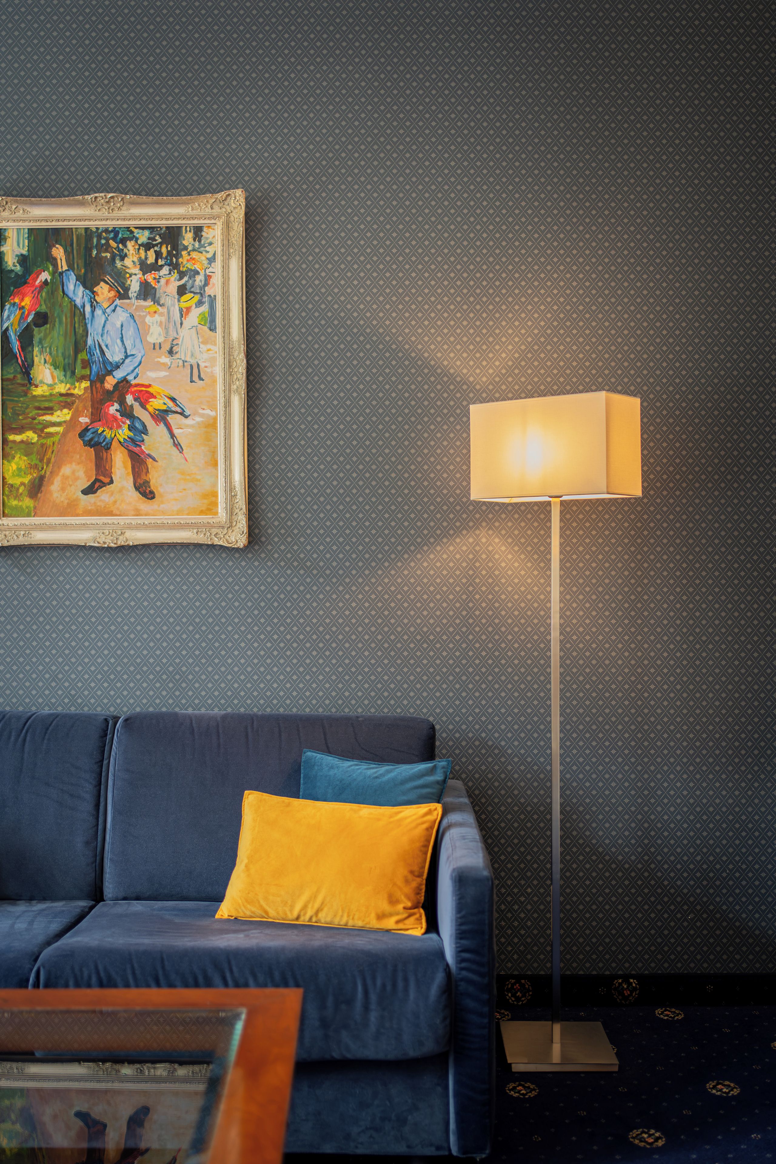A room with a dark patterned wall, a blue sofa with a yellow pillow, a framed painting, and a floor lamp.