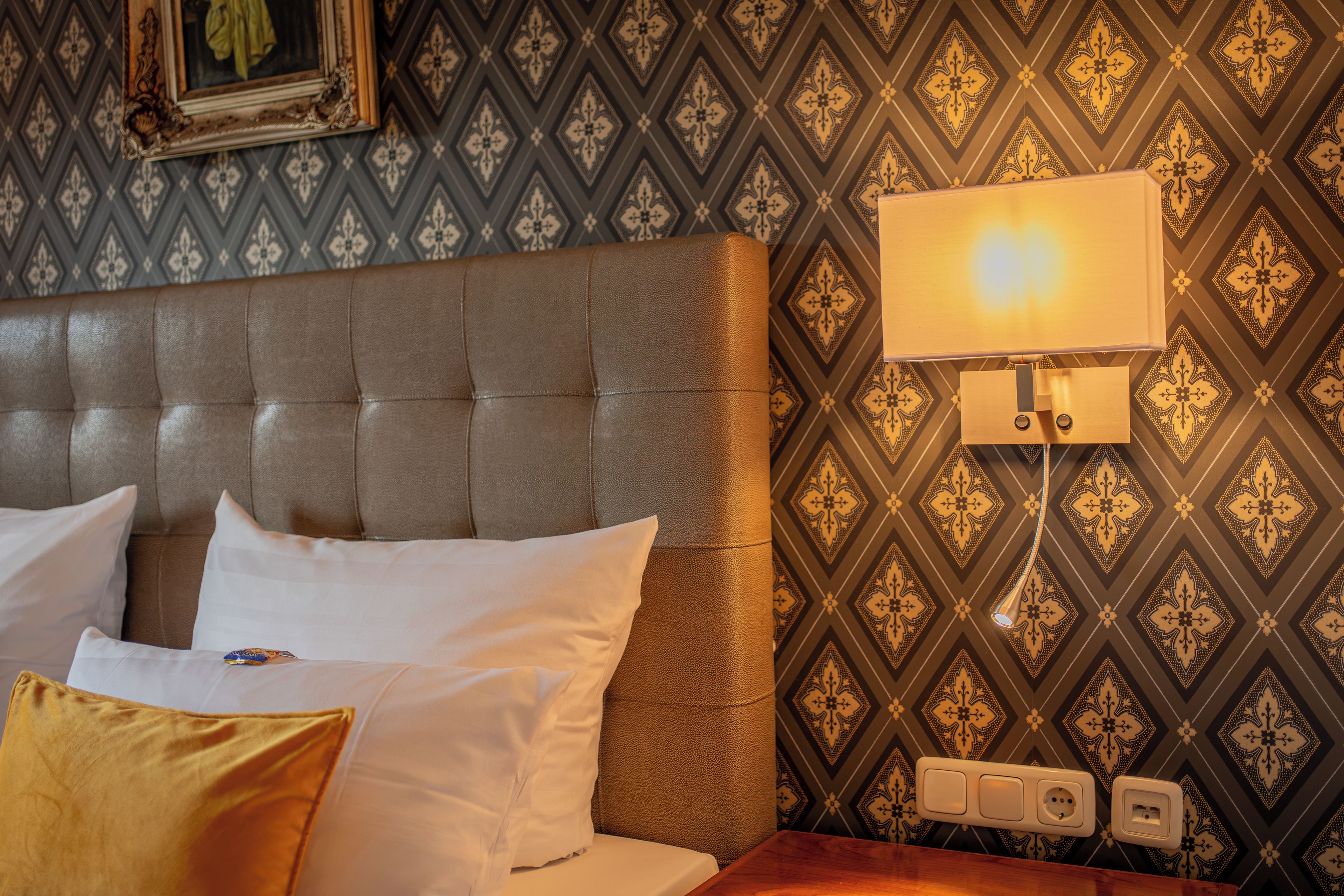 A hotel room with a tufted headboard, a bed with white and yellow pillows, a patterned wallpaper, and a bedside table with a lamp.