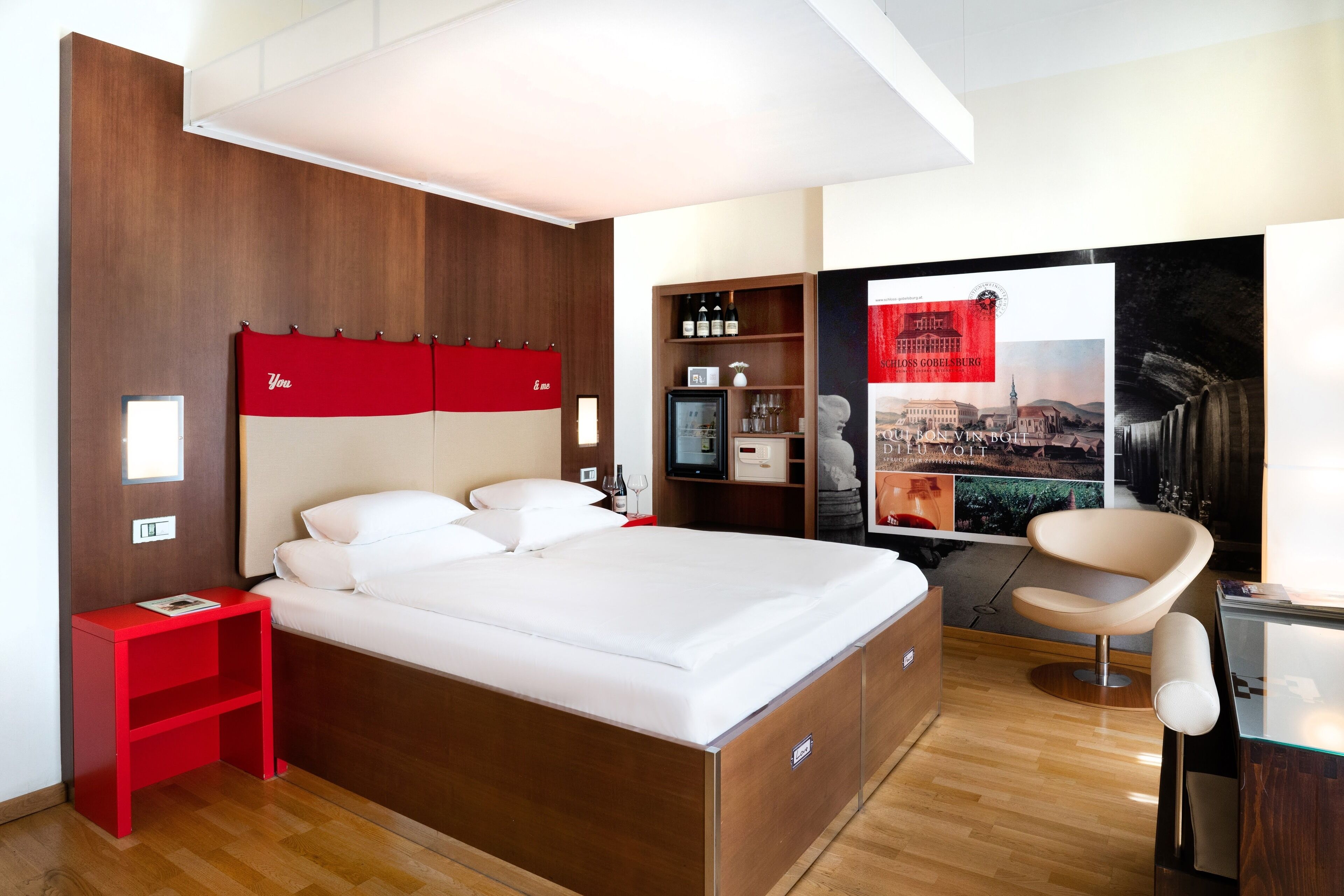 Une chambre d’hôtel moderne avec une tête de lit en bois, une table de chevet rouge, un lit avec du linge de maison blanc, un coin salon avec un fauteuil et une vue sur le paysage urbain par une grande fenêtre.
