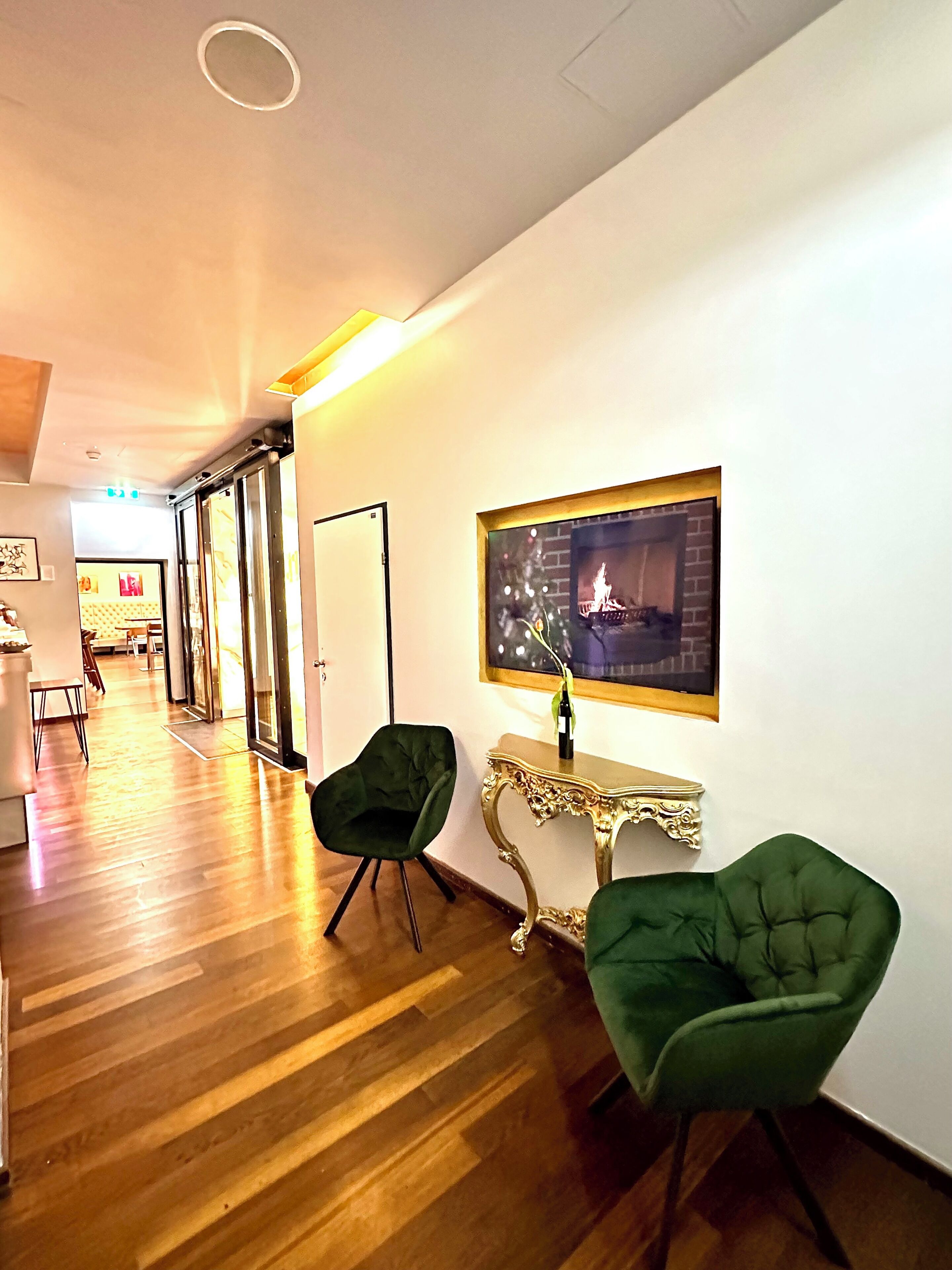 A hallway with wooden flooring, two green chairs, a gold-framed console table with a bottle, and a framed picture of a fireplace.