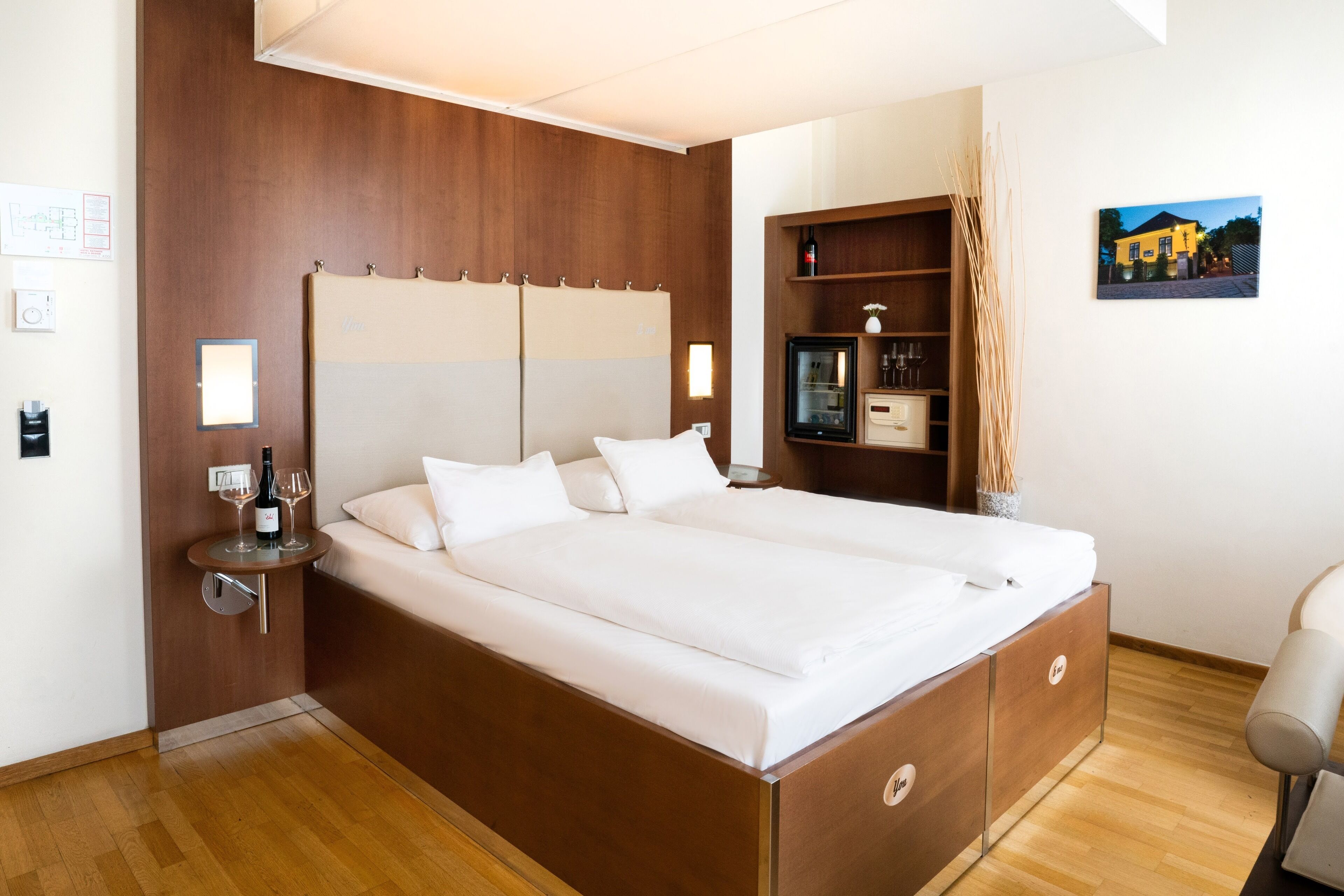 A modern hotel room with a large bed, wooden headboard, bedside tables, and a small side table with wine glasses.