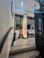 Um saguão de hotel com balcão de check-in, mesa de recepção e escada para os andares superiores.