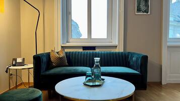 Una sala de estar con un sofá verde, una mesa redonda con una jarra de vidrio, una lámpara y un teléfono sobre una mesita auxiliar.