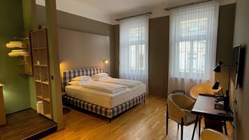 A hotel room with a bed, a desk, and a chair. There is a mirror, a shelf with towels, and a window with curtains.