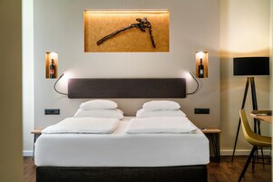 Quarto de hotel moderno com uma cama grande, mesas de cabeceira, escrivaninha e uma cadeira.