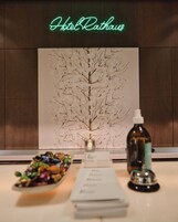 Uma área de recepção de hotel com um letreiro de neon que diz "HotelRathaus", uma obra de arte com tema de árvore emoldurada, um pequeno vaso com uma vela, um frasco de álcool em gel e uma pilha de enfeites coloridos.