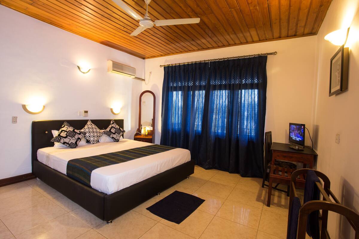 A bedroom with a bed, a desk, a chair, a ceiling fan, and a TV.