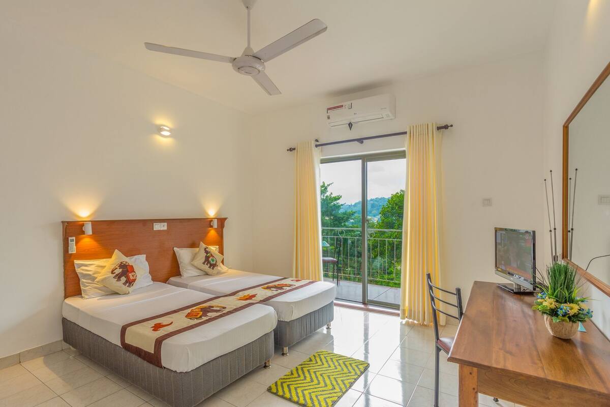 A hotel room with two beds, a ceiling fan, an air conditioning unit, a balcony with a view, and a dining table with a plant.