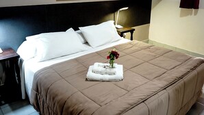 Una cama bien hecha con una colcha marrón, almohadas blancas y un pequeño florero con flores rojas sobre una toalla doblada.