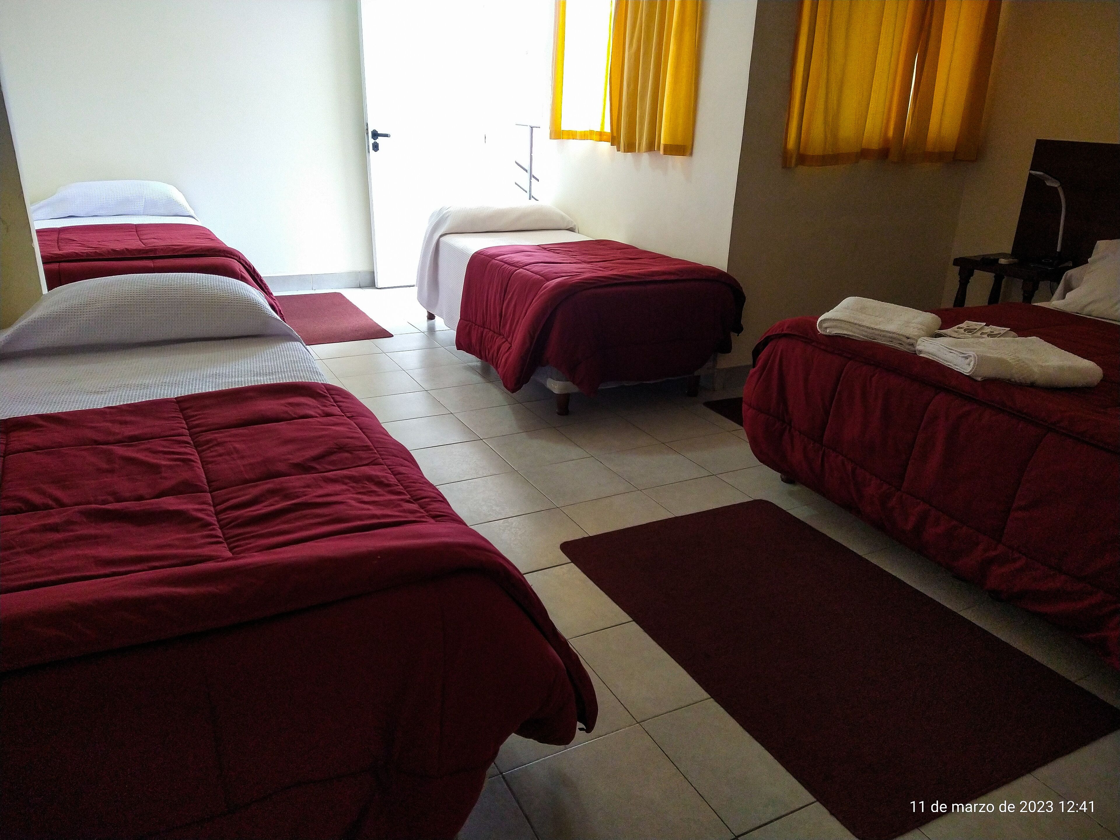 A hotel room with three beds, each with a red bedspread and white pillows. There is a small table with a lamp and a chair, and a window with yellow curtains.