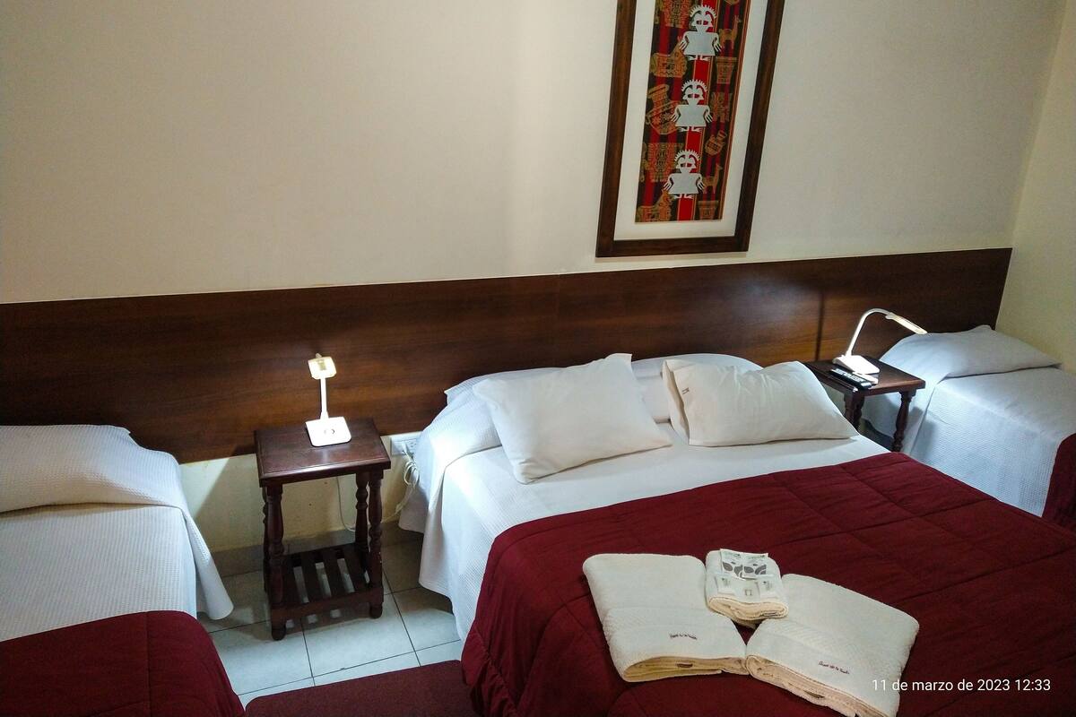 A hotel room with two beds, a nightstand with a lamp, a framed picture on the wall, and a red bedspread.