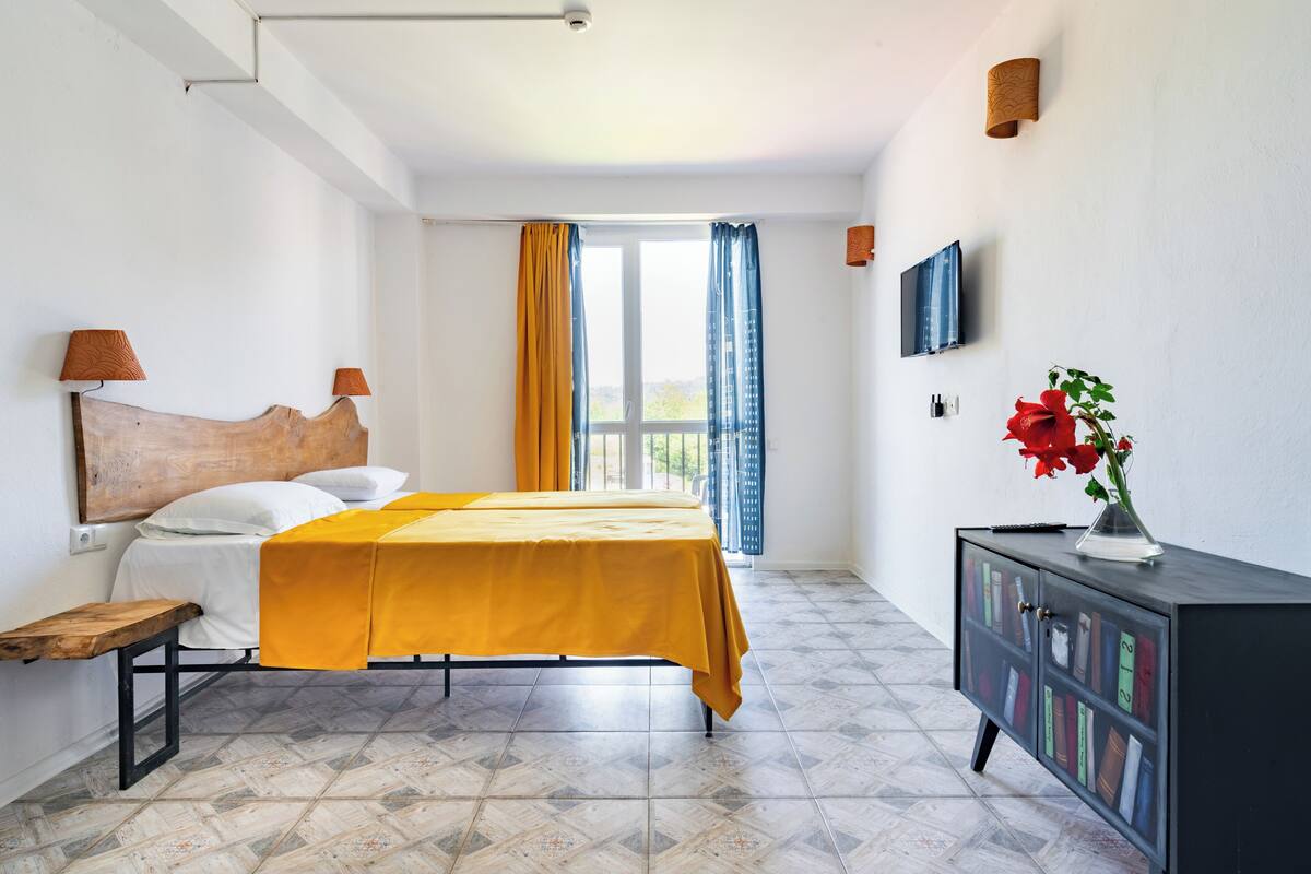 Ein Schlafzimmer mit einem Bett, einem Nachttisch, einer Kommode mit Büchern, einem Fernseher und einem Fenster mit Vorhängen.