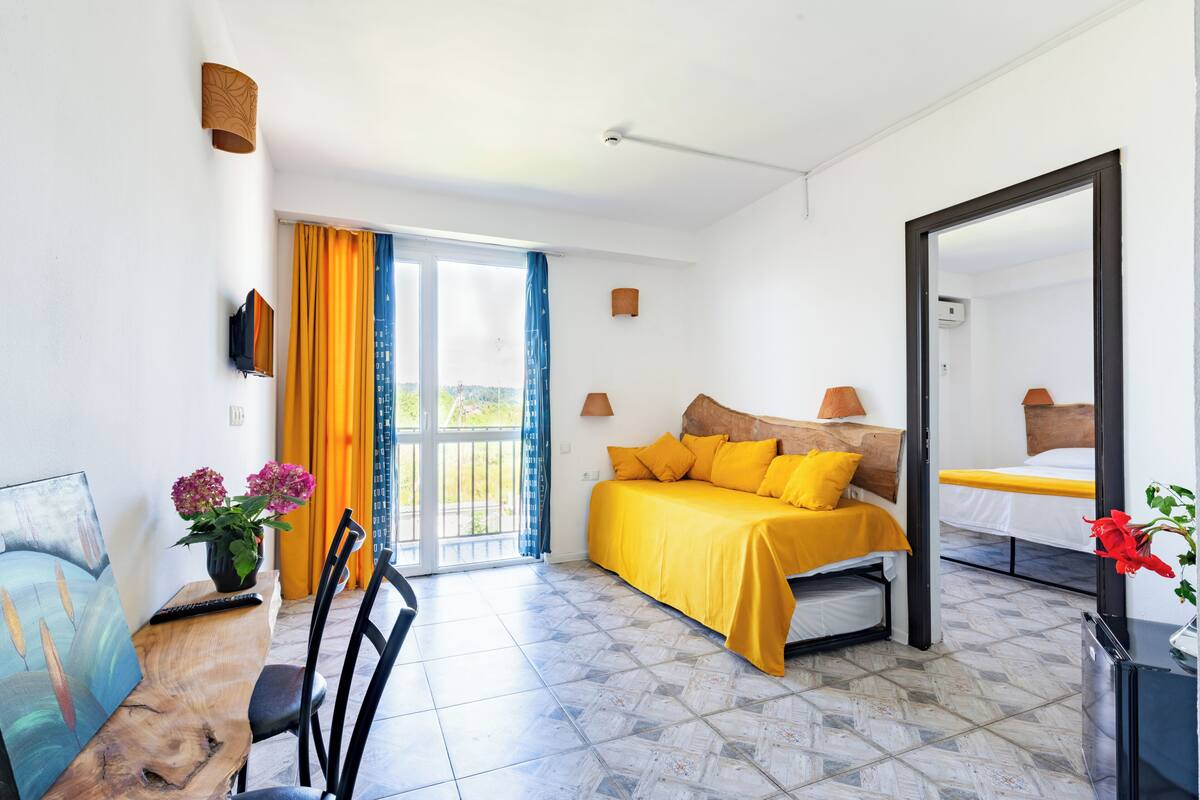 Ein Schlafzimmer mit einem Bett, einem Schreibtisch mit Stuhl, einem großen Fenster mit Vorhängen und einem Spiegel.