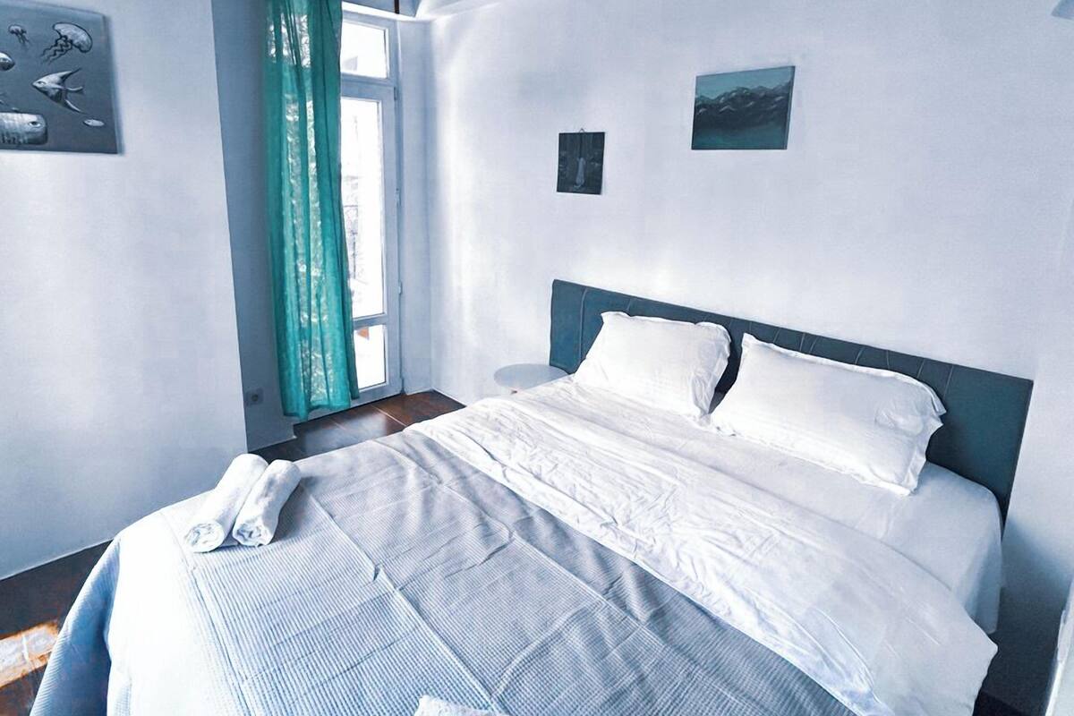 Ein Schlafzimmer mit Holzboden, einem Bett mit weißer Bettwäsche, einem Fenster mit Vorhängen und einem an der Wand hängenden Kunstwerk.