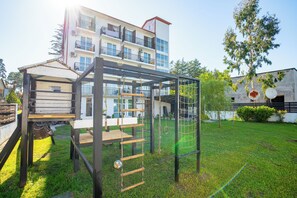 A playground with a climbing structure and swings, set against a building with balconies and a clear sky. - Simple Pleasures Shekvetili Hotel (Ureki)